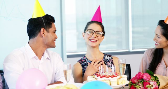 Smiling colleagues wearing party hats celebrating a birthday in modern office. Scene includes birthday cake and decorations, showing a friendly and joyous corporate environment. Ideal for use in articles or advertising about positive office culture, team bonding activities, or corporate events.