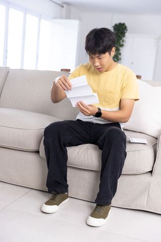 Asian man sitting on sofa reading letter with surprised expression in modern living room