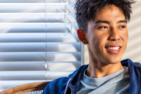 Sunlit asian man in 20s relaxing in rattan chair, window blinds casting light stripes