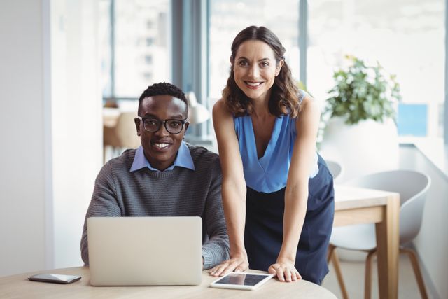 Diverse executives smiling while using a laptop in a modern office setting. Ideal for corporate websites, business presentations, marketing materials, or any content related to teamwork, collaboration, and professional work environments.