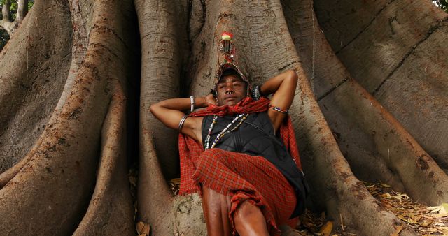 Indigenous man in traditional attire relaxing against enormous tree roots. Perfect for use in cultural studies, nature, relaxation, and travel-related content.