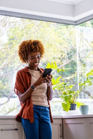 African american woman laughing while using smartphone in bright kitchen with plants