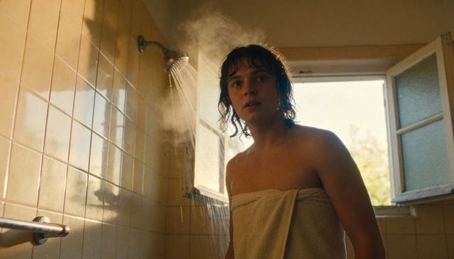 Young man wrapped in towel standing under steaming shower by open window, warm light