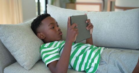 African american boy reclining on sofa using tablet, cozy home leisure and screen focus