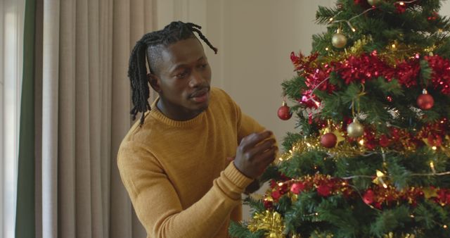 Happy african american man with dreadlocks decorating christmas tree, slow motion. Christmas, tradition, celebration, domestic life and lifestyle, unaltered.