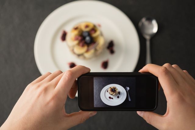 The image shows a person taking a photo of a plated breakfast using a smartphone. The top-down view highlights the food and the technology used to capture it. Ideal for illustrating concepts of food photography, social media sharing, modern dining experiences, and the intersection of culinary arts with technology.