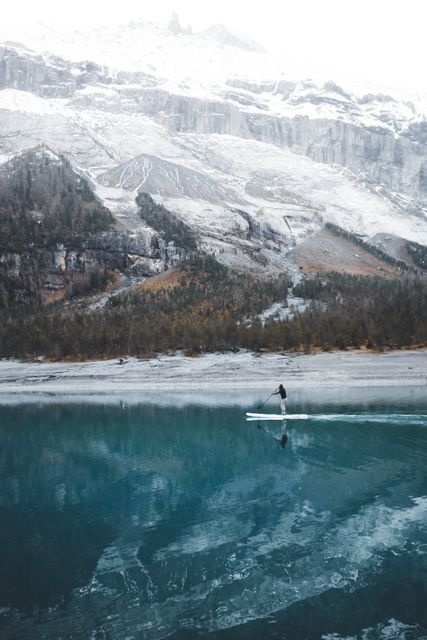 This photo shows an individual paddleboarding on a calm mountain lake, which beautifully reflects the snowy peaks. Ideal for travel, nature, and adventure blogs. Useful for promoting outdoor sports, winter activities, and serene nature retreats.
