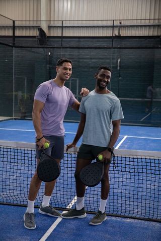Smiling diverse teammates on blue padel court holding paddles and yellow balls in casual sportswear