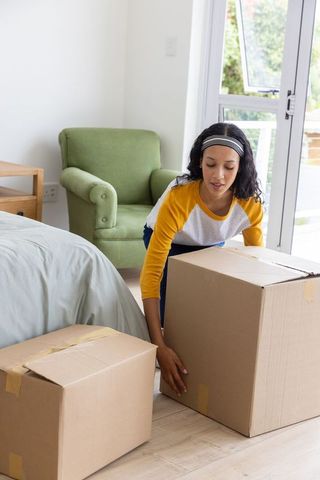 Teenage girl lifting moving boxes in bright minimal bedroom with green armchair