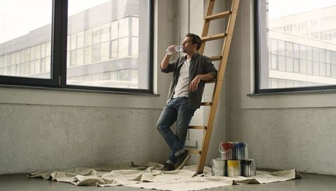 Man leaning on ladder drinking water during loft renovation with paint cans and drop cloth
