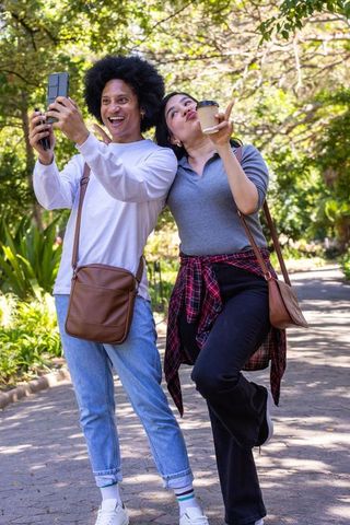 Young diverse couple posing for selfie on sunlit park path holding coffee and laughing