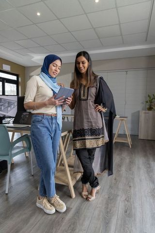 Two female colleagues wearing headscarf and tunic reviewing tablet in modern design studio