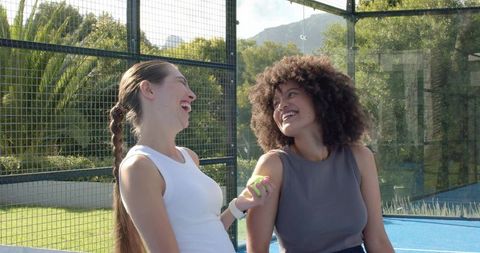 Women laughing on sunny outdoor sports court holding tennis ball, friendship and fitness