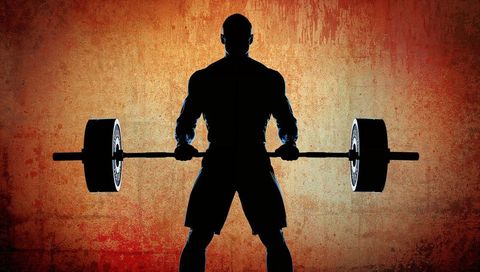 Backlit silhouette of muscular weightlifter holding heavy barbell against grunge wall