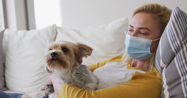 Woman sits on a couch wearing a face mask, holding her dog. Suitable for topics related to indoor lifestyle, home comfort, pet companionship, pandemic safety measures, or health and wellness.