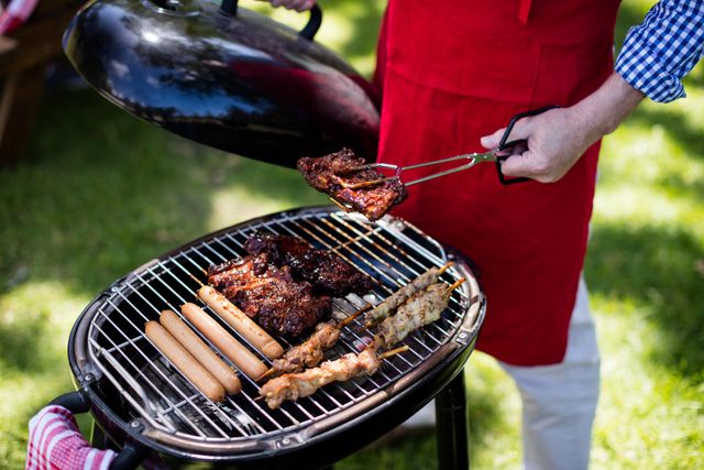 Man grilling various meats and sausages on a charcoal grill in a park. Ideal for use in content related to outdoor cooking, summer activities, family gatherings, and lifestyle blogs. Perfect for illustrating articles about barbecue recipes, picnic ideas, and weekend leisure activities.