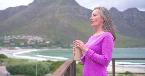 Middle-aged woman standing on coastal balcony holding bottle gazing bay and mountains