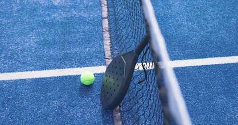 Padel paddle leaning on net with neon ball on textured blue court closeup