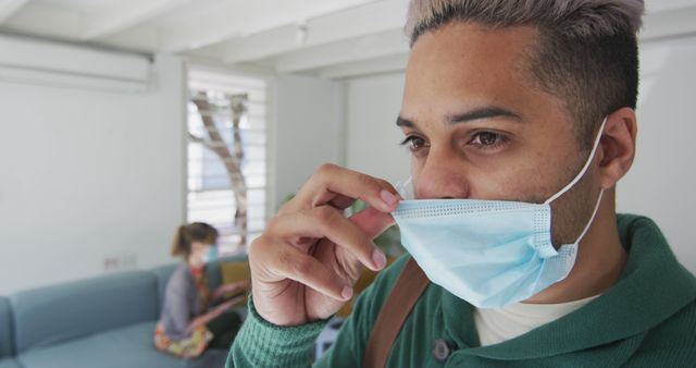 Man in indoor setting adjusting face mask, indicative of safety measures during pandemic. Suitable for health advisories, safety guidelines, COVID-19 awareness materials, and public health campaigns.