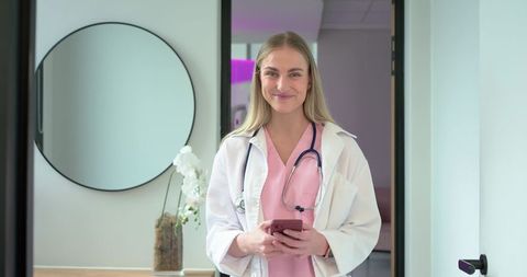Smiling female clinician standing in modern clinic holding smartphone wearing pink scrubs
