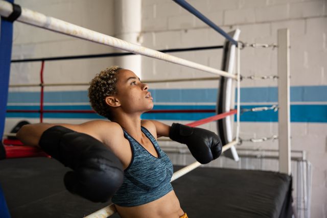 Biracial female boxer practicing in a boxing gym wearing sports clothes, resting leaning on ropes of a boxing ring. Strength sports achievement.