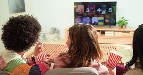 Diverse friends watching streaming service on cozy sofa holding red white striped popcorn