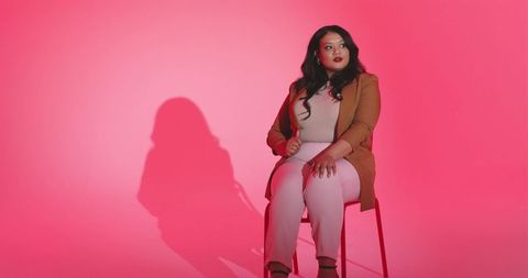 Confident mid adult woman sitting in blazer against pink gradient backdrop casting shadow