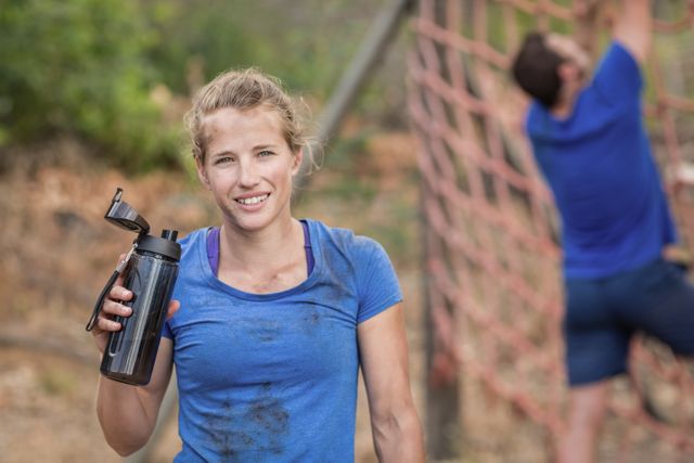 Woman holding water bottle during outdoor obstacle course training. She is wearing a blue shirt and appears to be taking a break. In the background, another person is climbing a rope net. Ideal for use in fitness, training, and outdoor activity promotions, as well as motivational and teamwork-related content.