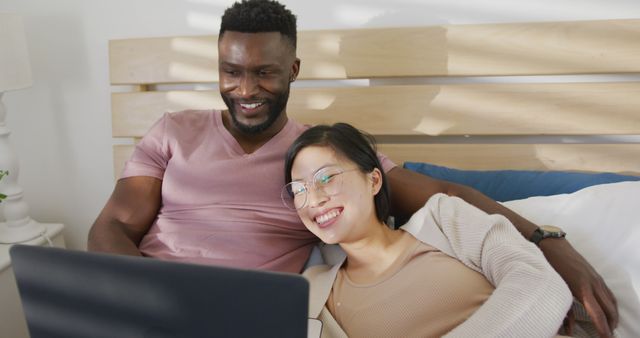 Happy diverse couple using laptop and lying in bedroom. Spending quality time at home concept.