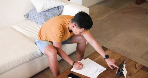 Man engaging with tablet while writing on documents at home