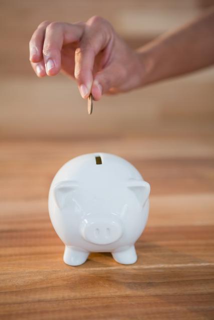 Hand inserting coin into white piggy bank on wooden surface. Ideal for illustrating concepts of saving money, financial planning, budgeting, and personal finance. Useful for blogs, financial advice websites, educational materials, and advertisements promoting savings and investments.