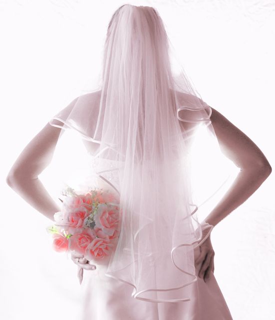 A bride is posing with her back to the camera, holding a bouquet of pink roses. She is dressed in a white bridal gown with a flowing veil. Ideal for wedding planning websites, bridal magazines, and wedding invitation designs.