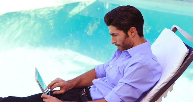 Young man in a blue shirt working on a laptop while lounging poolside. Ideal for illustrating concepts of remote work, outdoor productivity, casual business attire, and a balanced work-life lifestyle.
