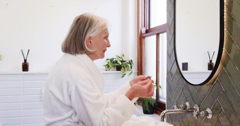 Senior woman in bathrobe enjoying morning skincare routine in bathroom