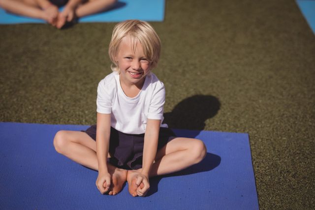 This image shows a young child smiling while practicing yoga on a blue mat outdoors. Ideal for use in educational materials, fitness programs for children, or promotional content for outdoor activities and healthy lifestyles.