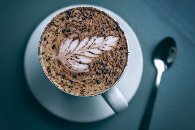 Close up of a cappuccino with intricate latte art, served on a dark gray table with spoon. Ideal for use in coffee shop advertisements, cafe menus, food blogs, social media promotions for coffee houses, or articles about coffee culture.