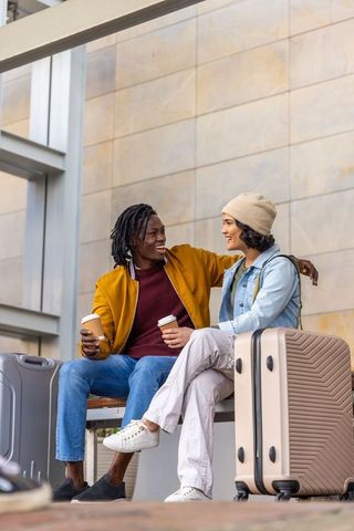 Diverse couple sitting at station concourse with luggage holding coffee cups and chatting