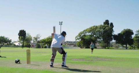 Cricketer driving shot while fielder sprinting to throw at stumps on sunny park pitch
