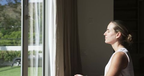 Woman basking in morning sun at sliding glass door meditating in minimalist room