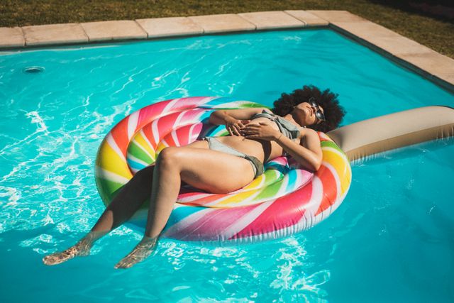 Biracial woman enjoying a sunny day while lying on a colorful inflatable toy in a swimming pool. Ideal for use in advertisements for summer vacations, pool accessories, swimwear, and outdoor leisure activities. Perfect for lifestyle blogs, travel websites, and promotional materials for resorts and hotels.