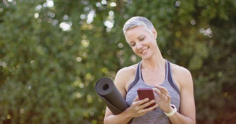 Senior woman checking smartphone after outdoor yoga session holding rolled mat smiling