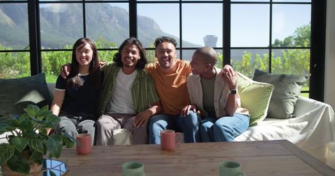 Diverse friends laughing together on sunlit living room sofa with coffee mugs