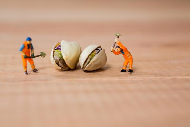 This image shows two miniature construction workers in orange uniforms using tools to break open pistachio nuts. It creatively combines elements of construction work with food, making it ideal for use in advertisements, social media posts, or articles related to creativity, teamwork, or food. The playful and humorous nature of the image can attract attention and add a unique touch to various projects.