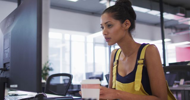 This stock photo depicts a biracial businesswoman working at her desk, deeply focused on her computer monitor. This image is perfect for illustrating concepts related to technology in the workplace, corporate environments, and professional multitasking. Use it in articles, blogs, or presentations about modern office aesthetics, career development, women in business, or the rise of independent creative enterprises.