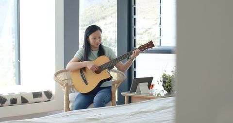 Asian woman playing acoustic guitar in sunlit bedroom with tablet for online lessons
