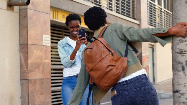 Two African American twin sisters enjoying time on the city sidewalk while taking a photo together. One sister is holding a smartphone and clicking a picture, while the other is striking a pose with a backpack. Ideal for themes of sibling bonds, city life, casual style, and youthful energy.