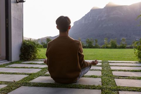 Man meditating on stone paver patio facing mountain ridge at sunlit dawn, lawn terrace