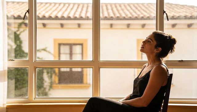 Sunlit woman sitting on chair gazing through paned window at tiled roof, minimalist room