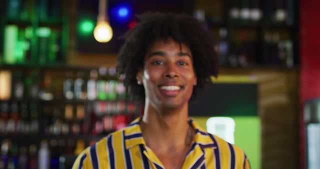 Man smiling while standing at a bar with colorful lights and bottles in the background. Suitable for themes related to joy, social events, bars, nightlife, and casual gatherings.