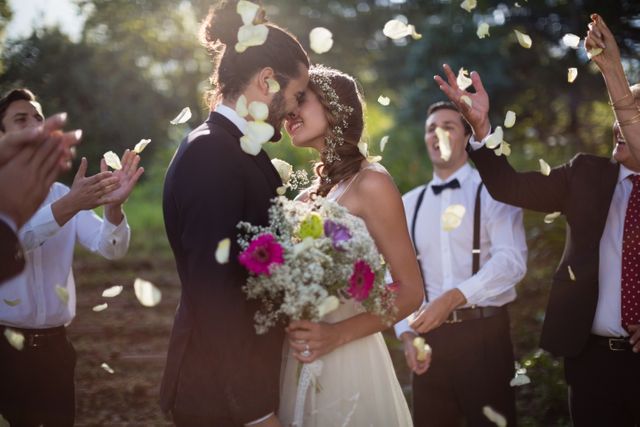 Bride and groom sharing a romantic kiss surrounded by friends throwing petals in a park. Ideal for wedding invitations, romantic greeting cards, wedding planning websites, and bridal magazines.